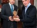 Dos hombres posan con un premio en forma de rueda de barco en un ambiente elegante con decoración clásica.