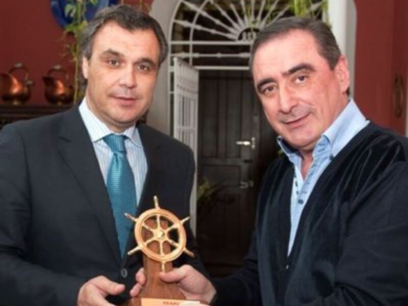 Dos hombres sostienen un trofeo en forma de rueda de timón, posiblemente en un evento de premiación o reconocimiento.