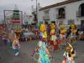 Un desfile de carros alegóricos con figuras coloridas y personajes en el centro, frente a una casa blanca con balcones. La imagen muestra un ambiente festivo y celebrativo, típico de una fiesta o carnaval.