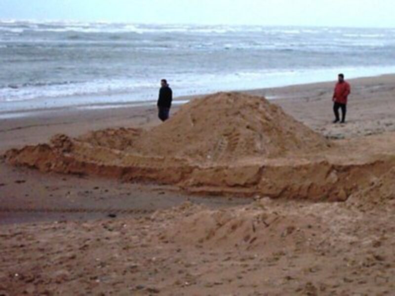 Un castillo de arena en la playa con dos personas observándolo, el mar en el fondo.