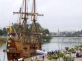 Una réplica de un velero colonial en el puerto, con gente observando y barcos en el fondo.