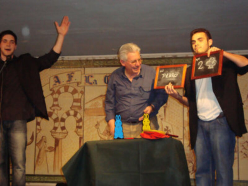 Tres personas en un escenario, una sosteniendo un tablero con '2' y otra con '1', posiblemente en un juego o presentación.