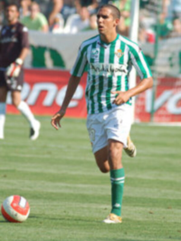 Jugador en acción durante un partido de fútbol, con uniforme verde y blanco, en un estadio con público.