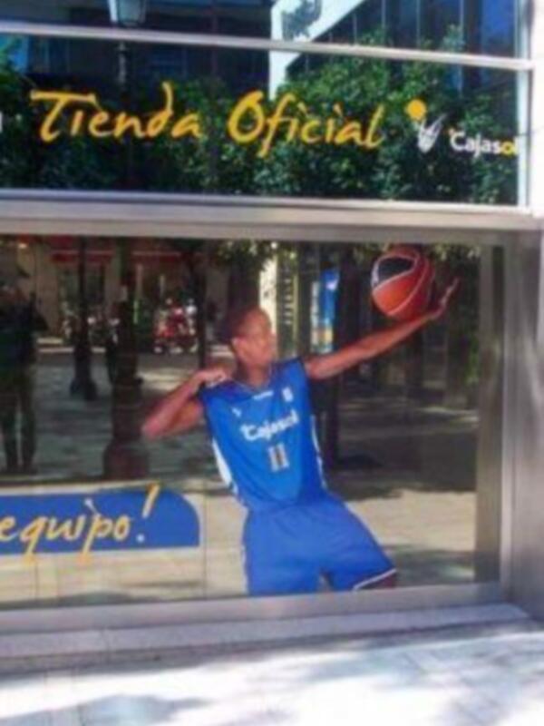 Un jugador de baloncesto en una camiseta azul con el logo de la marca 'Cajasol' en una ventana de tienda. El texto en la ventana dice 'Tienda Oficial Cajasol'.