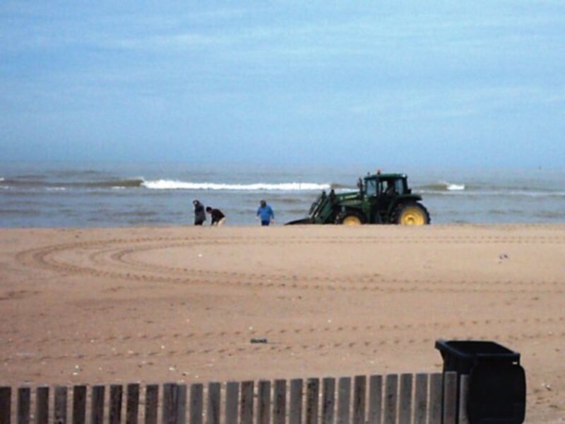 Tractor y personas en la playa, con rastros de ruedas y contenedores de basura al frente.