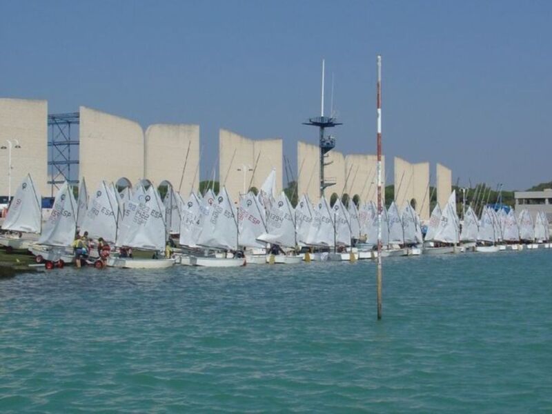 Un grupo de barcos de vela con banderas españolas en el agua, con un edificio moderno y una torre de control al fondo.