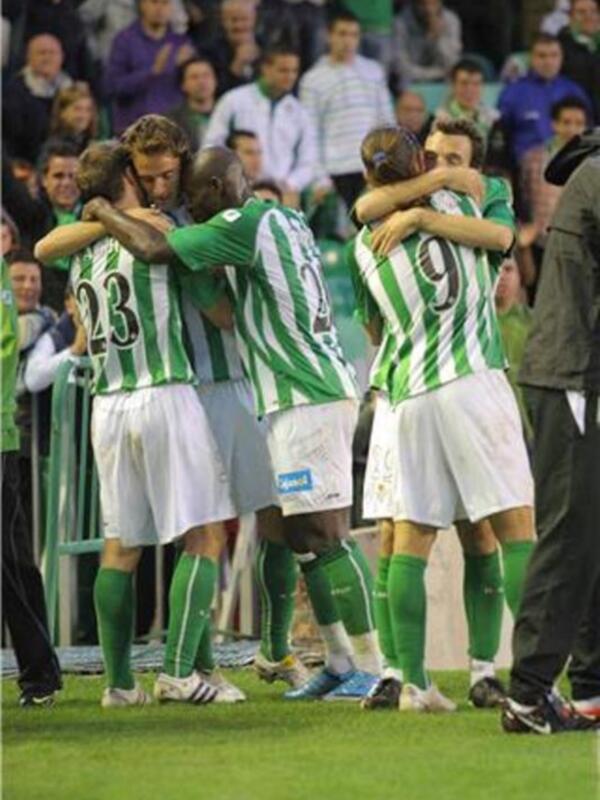 Un grupo de jugadores de fútbol celebrándose en el campo de juego, con una camiseta verde y blanca y una pelota verde en el suelo.