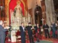 Una imagen de una ceremonia religiosa en un templo, con personas vestidas formalmente celebrando. En el fondo, una imagen de la Virgen María y un altar con velas encendidas. La decoración interior es elegante, con detalles dorados y rojos.