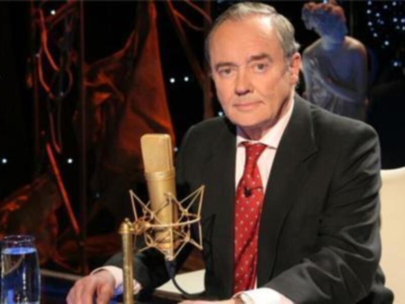 Presentador en un estudio de radio o televisión, vestido formalmente con un micrófono en frente.
