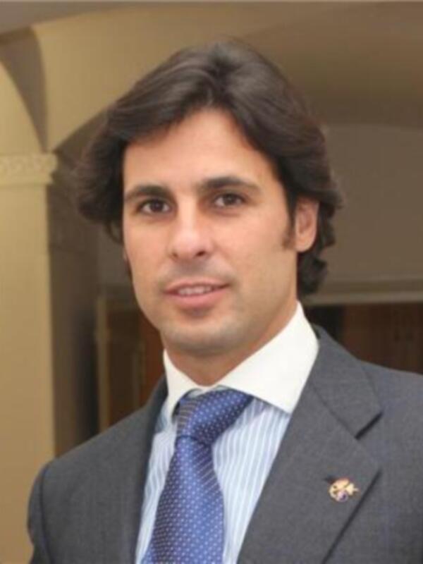 Retrato de hombre con traje y corbata, posando en un entorno interior con arcos y columnas.