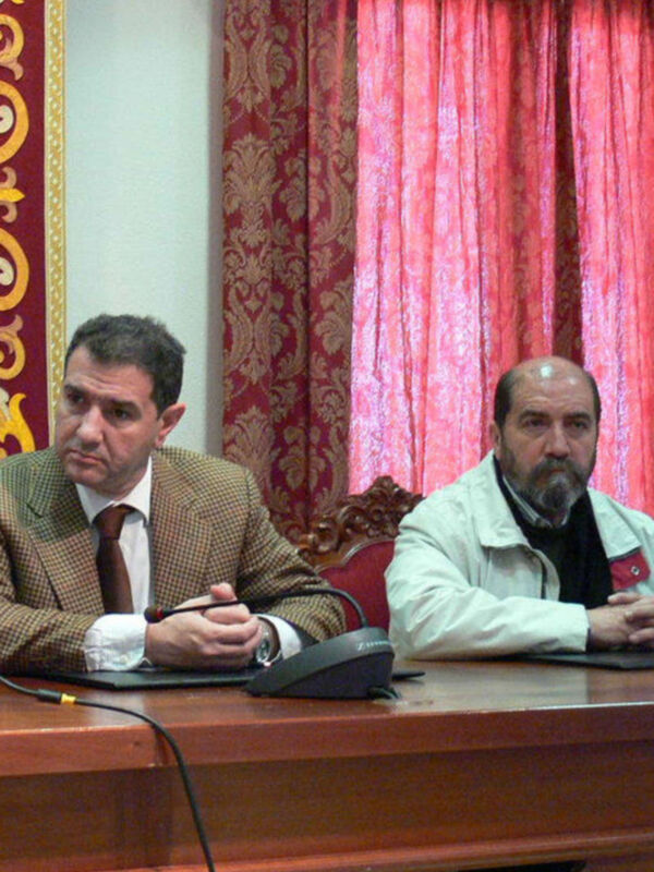 Dos hombres sentados en una sala con cortinas rojas, uno con traje y otro con chaqueta blanca, en una reunión o conferencia.