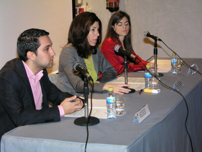 Panel de discusión con tres participantes sentados en una mesa con micrófonos y documentos.