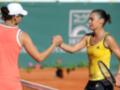 Dos jugadoras de tenis se saludan con una sonrisa, uno con un vestido naranja y la otra con un jersey amarillo. La superficie es de tierra batida, y el fondo muestra una publicidad de Roland Garros.