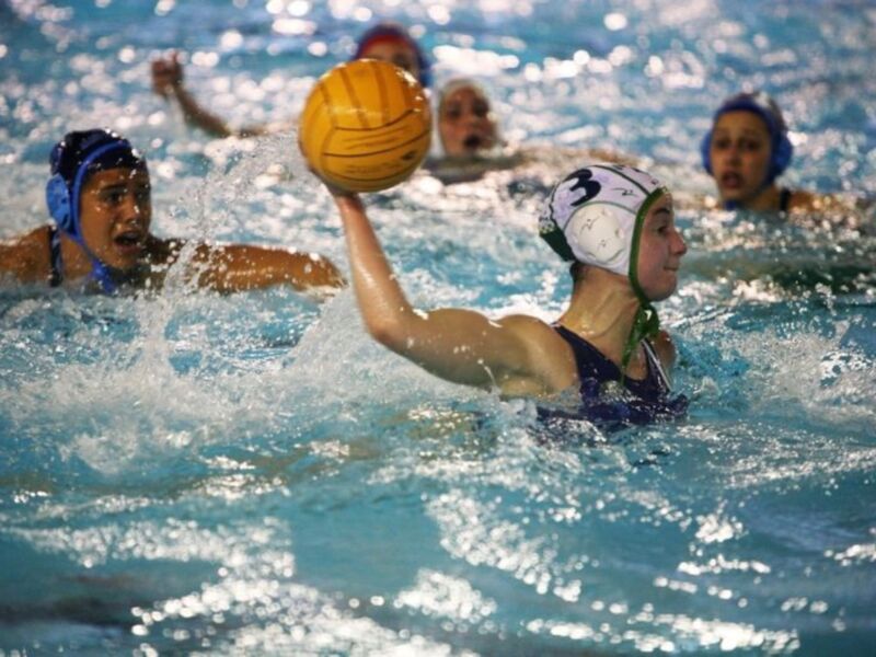 Una jugadora de waterpolo lanza la pelota hacia otro jugador mientras nadan en un estadio de natación.