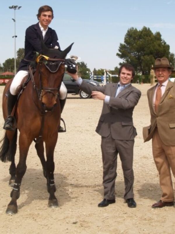 Joven jinete montando un caballo en un evento ecuestre, acompañado por dos hombres en trajes.