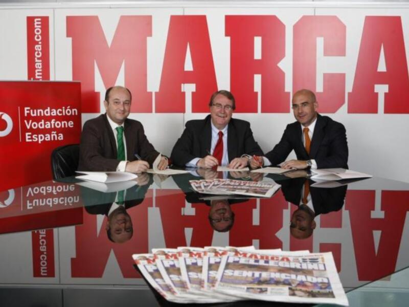 Tres hombres firmando documentos en una oficina de Fundación Marca, con el logo de Vodafone España en el fondo.