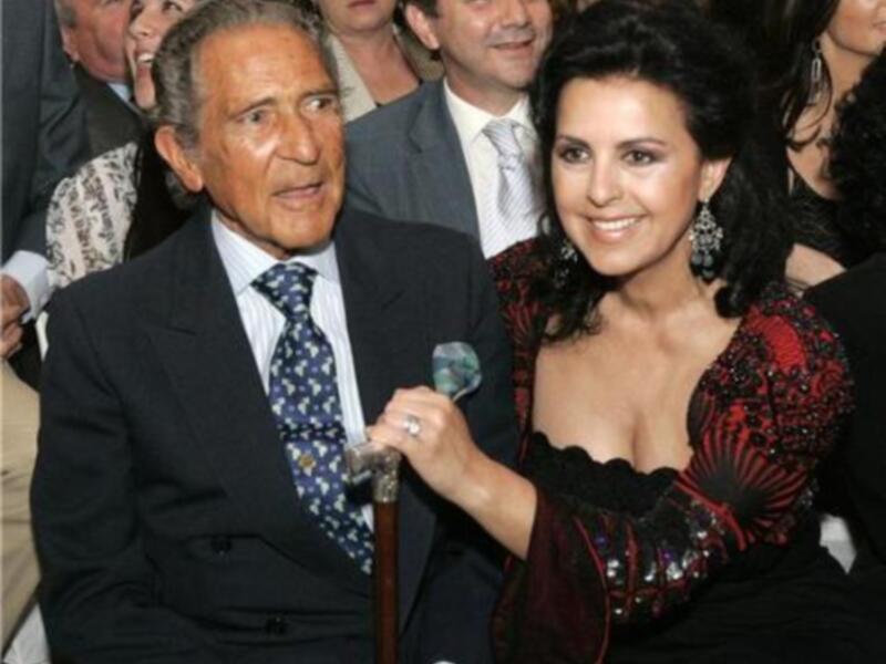 Una imagen de una mujer con un vestido negro y rojo brillante, posando junto a un hombre en un evento formal. La mujer sostiene un bastón y sonríe. La escena parece ser una celebración o una reunión social.