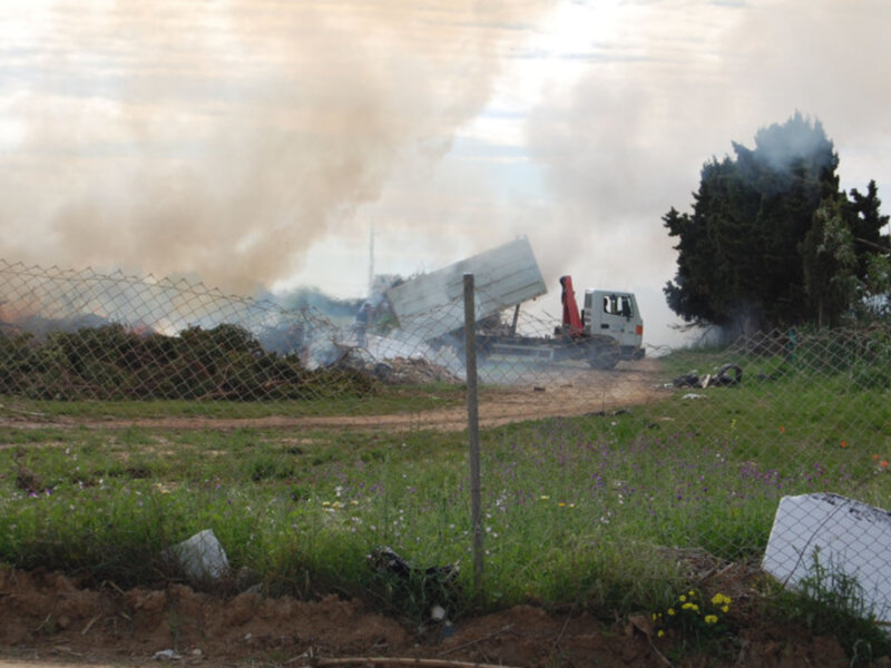 Vehículo de construcción en un campo con humo en el cielo, cerca de una cerca y arbustos.