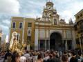 Una multitud de personas se concentra en una plaza frente a una iglesia monumental, con detalles dorados y un arco principal. La imagen transmite la celebración de una festividad religiosa, con estatuas doradas y decoraciones navideñas.
