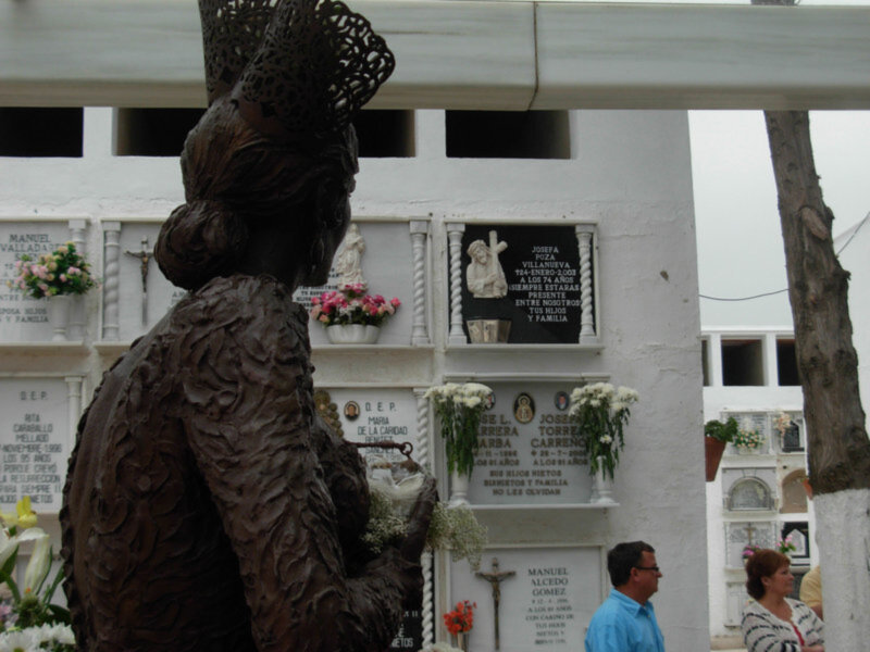Estatua de una mujer con mantón de color en un cementerio blanco, con detalles de flores y lápidas en el fondo.