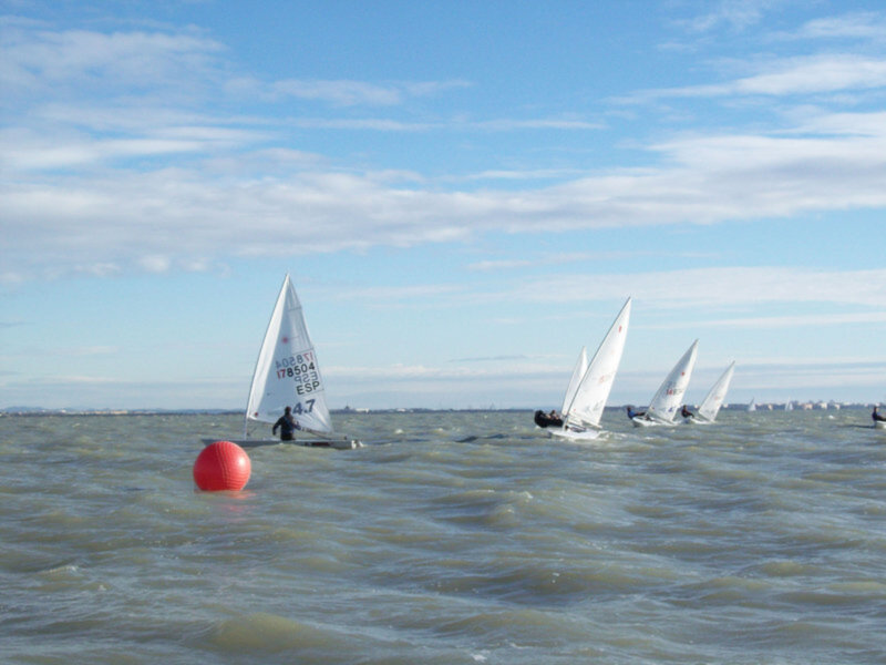 Sailboats on the water with a red buoy in the foreground and a blue sky with clouds in the background.