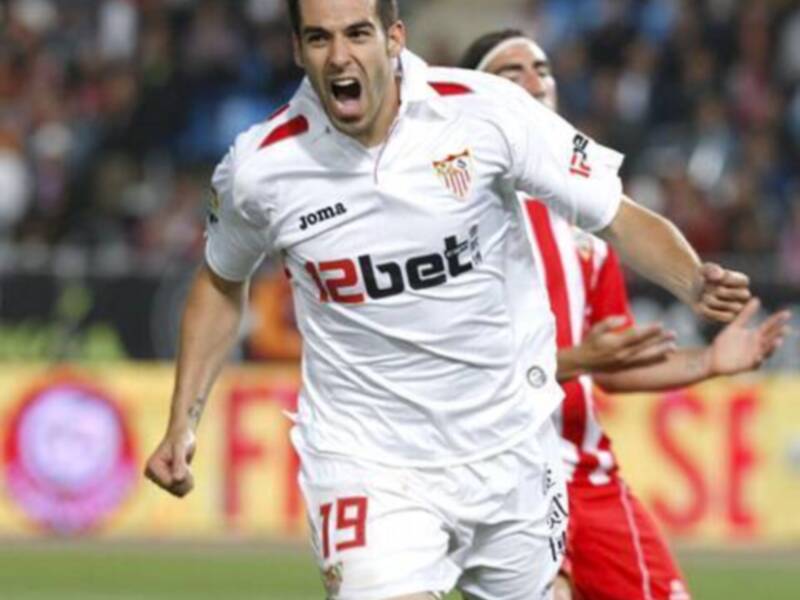 Un jugador de fútbol celebrando un gol en un partido de fútbol, con el escudo del Sevilla FC en su camiseta.