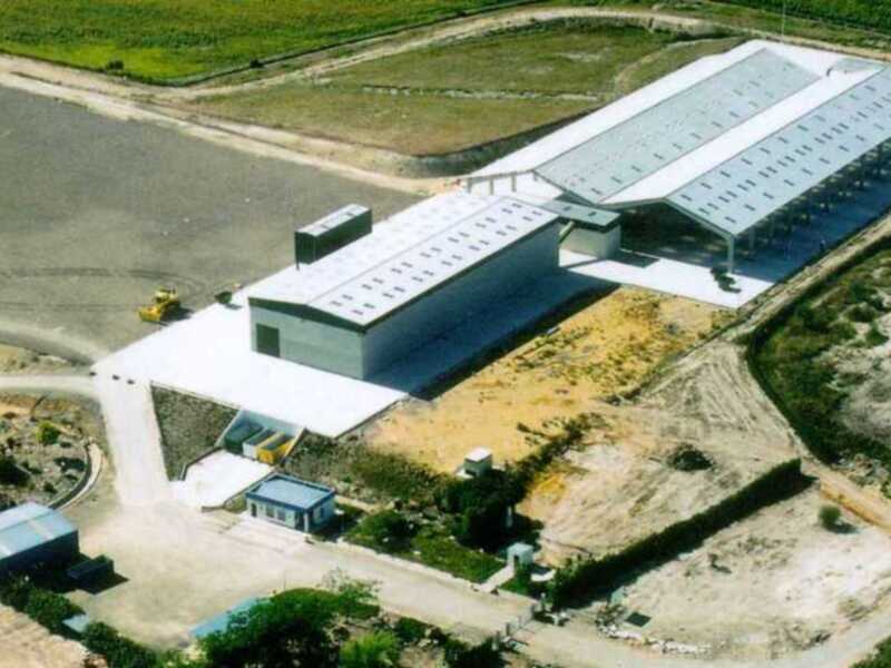 Fotografía aérea de una instalación industrial con un edificio principal y otros edificios adjuntos, ubicada en un área de tierras agrícolas.