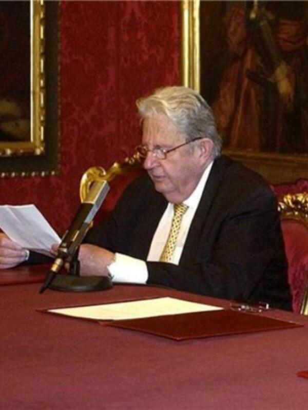 Persona en traje y corbata leyendo documentos en un ambiente formal con tapicería roja y marcos dorados.