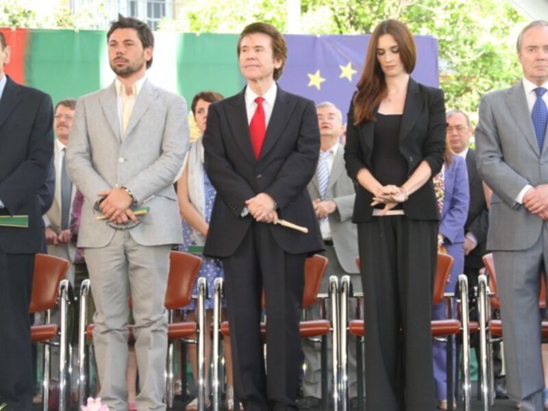 Persona en traje rojo y otros en trajes formales en un evento público al aire libre.