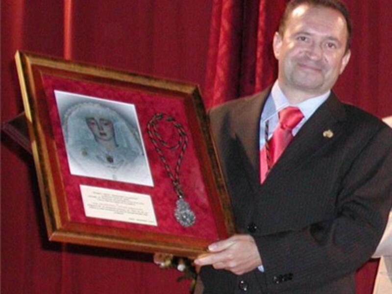 Persona en traje formal sosteniendo un marco con una medalla y una imagen de una figura religiosa.