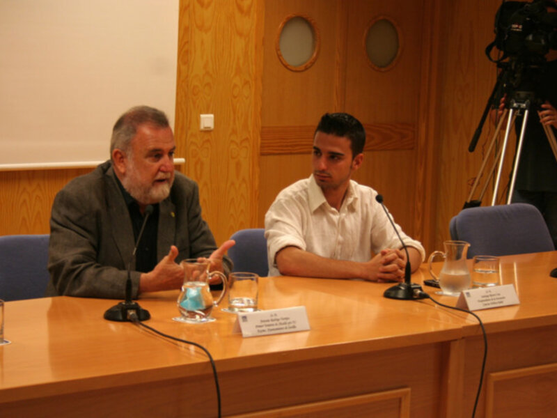 Dos hombres en una mesa de conferencia discuten mientras se graba con una cámara de video en el fondo.