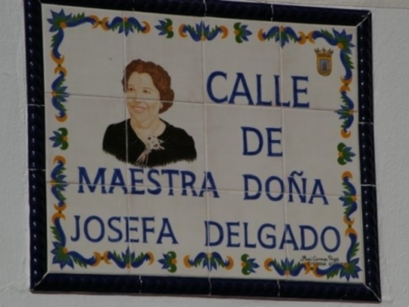 Placa conmemorativa dedicada a la Maestra Doña Josefa Delgado, decorada con motivos florales y un retrato en el centro.