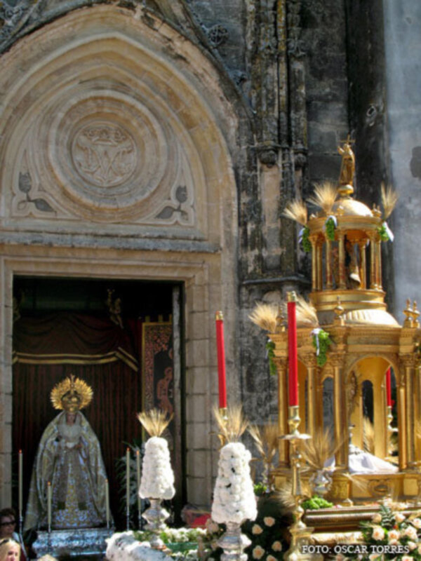 Procesión religiosa con estandartes y imagen de la Virgen en la plaza de una iglesia histórica.