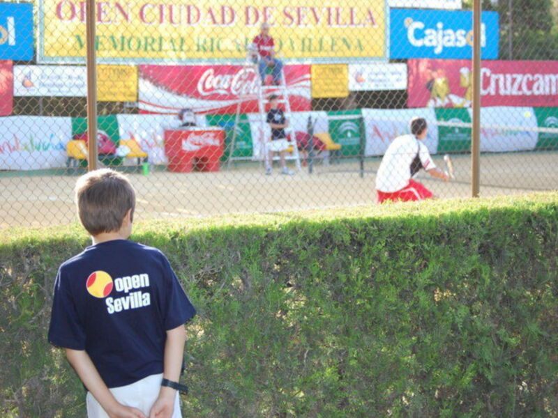 Un niño observa un partido de tenis en el Open Ciudad de Sevilla, con publicidad en el fondo y un jugador en acción en el campo.