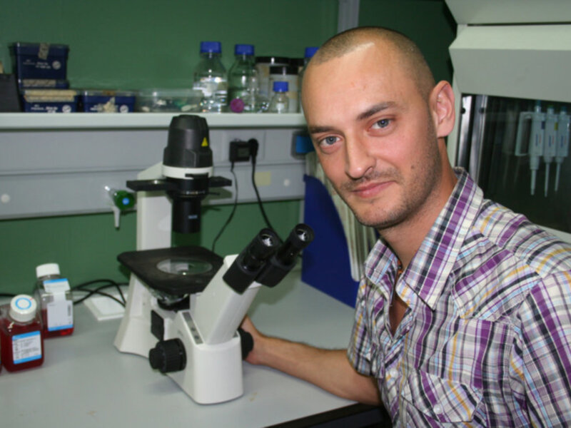 Un científico en un laboratorio, con un microscopio en primer plano y botellas de reactivos en el fondo.