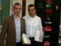 Dos hombres posan con folletos en un evento de la Fundación del Deporte de Andalucía, junto a logos de patrocinadores como Canal Sur y Cruzcampo.