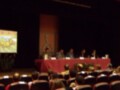 Conferencia en un escenario con panelistas sentados detrás de una mesa roja, frente a una pantalla que muestra un dibujo animado.