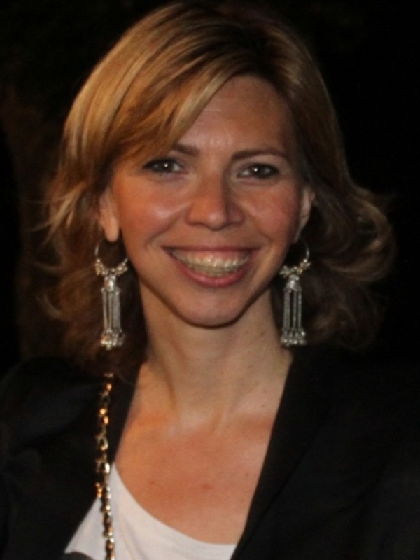 Una mujer con cabello largo y ondulado, llevando una chaqueta negra y un collar con cuentas de cristal. El fondo es oscuro, enfocándose en su rostro y parte de su vestimenta.