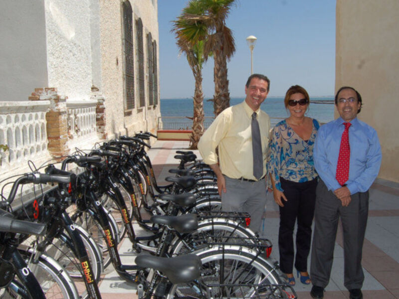 Tres personas posan junto a una fila de bicicletas en un entorno urbano con palmeras y mar al fondo.