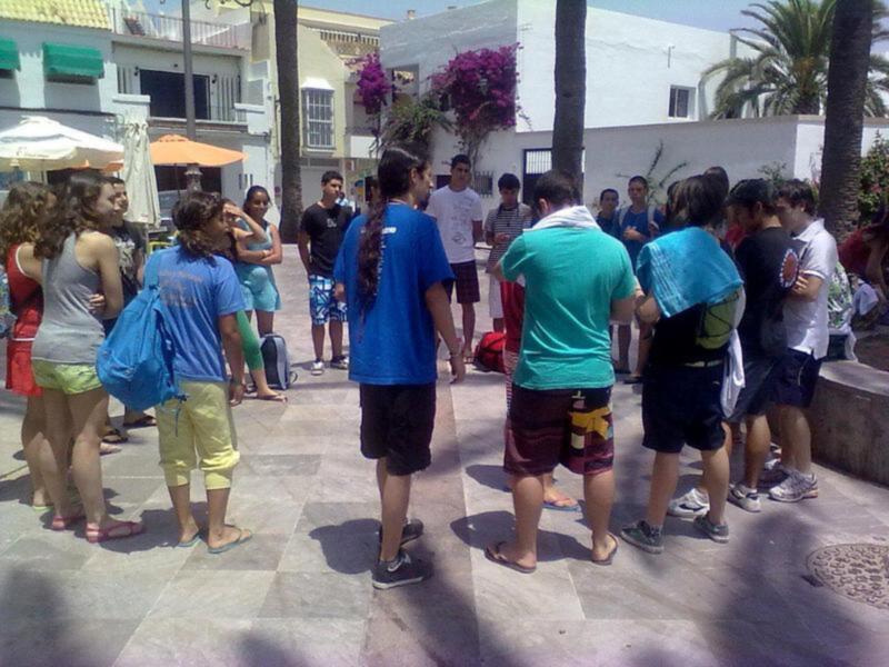 Un grupo de jóvenes se reúne en una plaza con palmeras y edificios blancos, posiblemente para una actividad o reunión al aire libre.