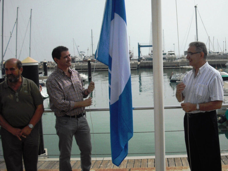 Celebración de una bandera frente al puerto con veleros en el fondo.