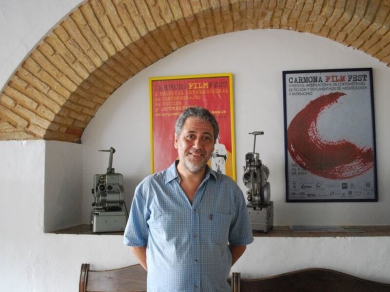 Un hombre sonriente frente a un cartel de cine en una sala con una máquina de café en el fondo.