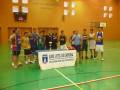 Equipo de baloncesto en un gimnasio municipal, con una pancarta que dice 'ILMO. AYTO. DE CHIPIONA DELEGACIÓN DE DEPORTES' y un reloj en el fondo que muestra la hora.