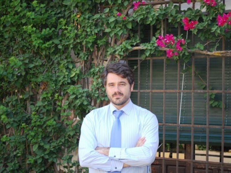 Hombre con camisa y corbata, brazos cruzados, frente a una pared con plantas y flores rosadas.