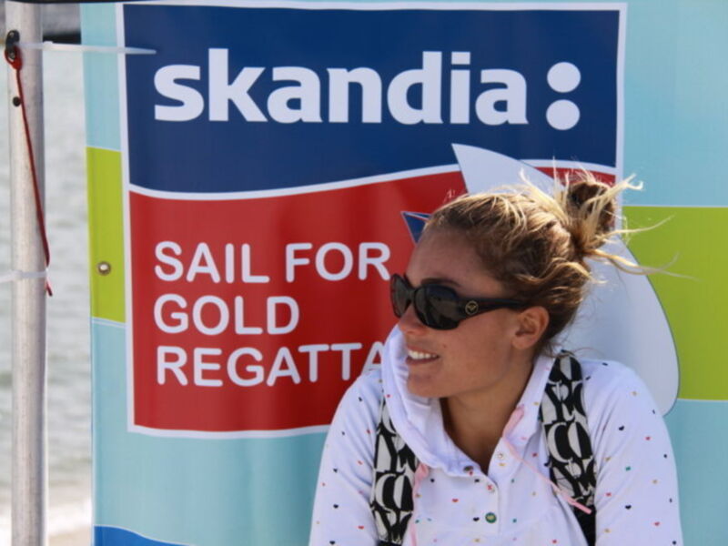 Una mujer sonríe mientras sostiene un surf cerca de un cartel que dice 'SAIL FOR GOLD REGATTA'.