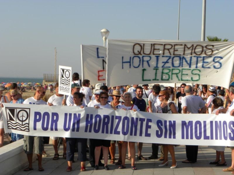Manifestación en la playa contra la instalación de molinos eólicos, con pancartas que dicen 'Por un horizonte libre sin molinos'.