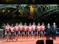 Equipo ciclista en ceremonia de presentación con puente iluminado de fondo.