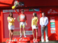 Un ciclista en el podio, con un maillot rojo y flores en la mano izquierda. Detrás de él, dos mujeres con vestidos amarillos y un hombre en traje blanco. El fondo es rojo con logos de sponsors.