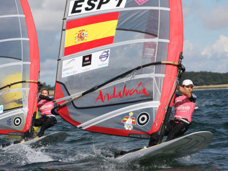 Competición de vela olímpica en Andalucía con vela española y deportistas en acción sobre el agua.