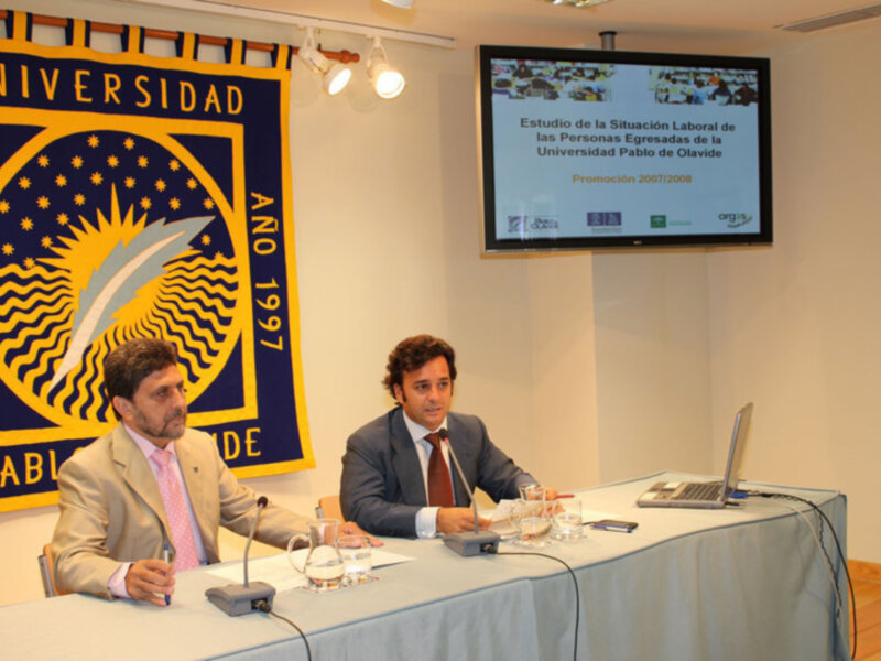 Dos hombres sentados en una mesa con un banner de la Universidad Pablo de Olavide en el fondo, presentando un estudio sobre la situación laboral de egresados de la promoción 2007/2008.